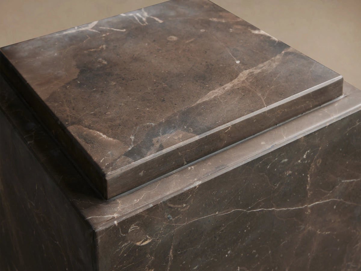Marmont Pedestal in dark natural marble with matte lacquer finish, featuring unique veining. Displayed on a wooden floor.