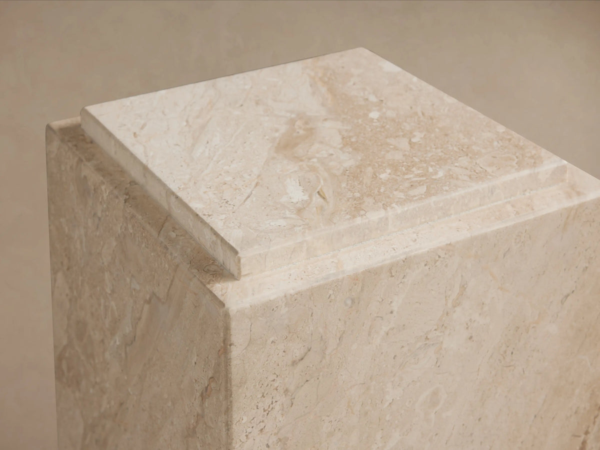 Marmont Pedestal made of beige natural marble with unique veining, matte lacquer finish; standing on a wooden floor against a beige wall.