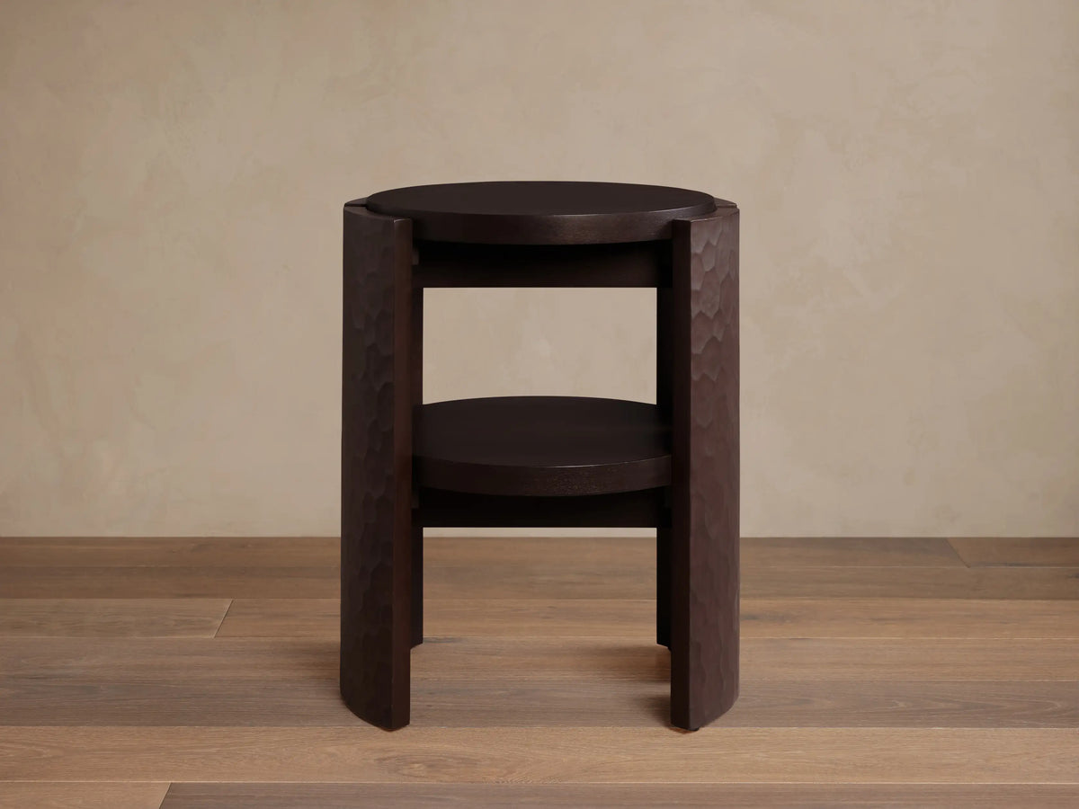 Dark brown Chiseled End Table with a round top and shelf, featuring deeply textured hand-carved legs, on a wood floor.