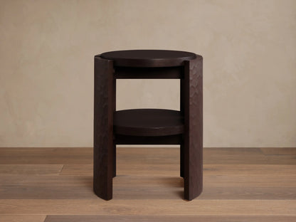 Dark brown Chiseled End Table with a round top and shelf, featuring deeply textured hand-carved legs, on a wood floor.