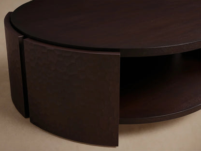 Dark mahogany chiseled round coffee table with textured curved side panels, round top, and lower shelf on wood floor.
