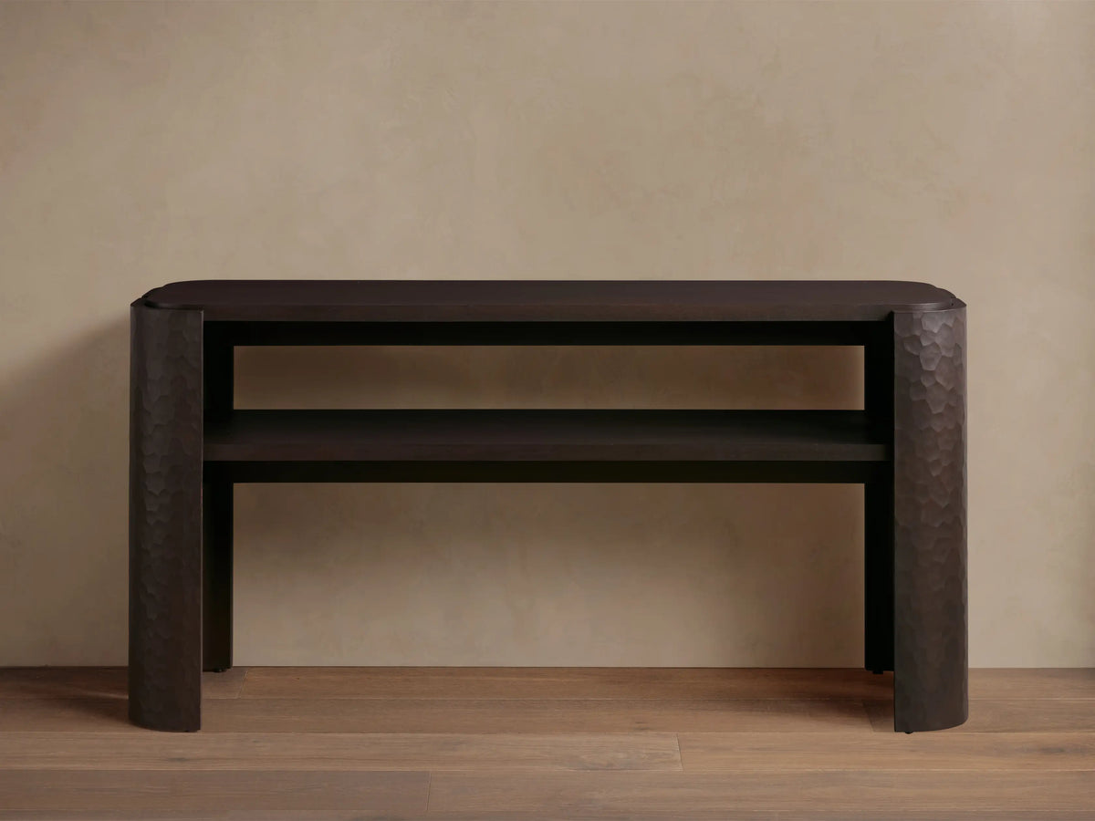 Dark brown mahogany Chiseled Console Table with rounded top, lower shelf. Legs feature distinct chiseled texture. Styled with decor.