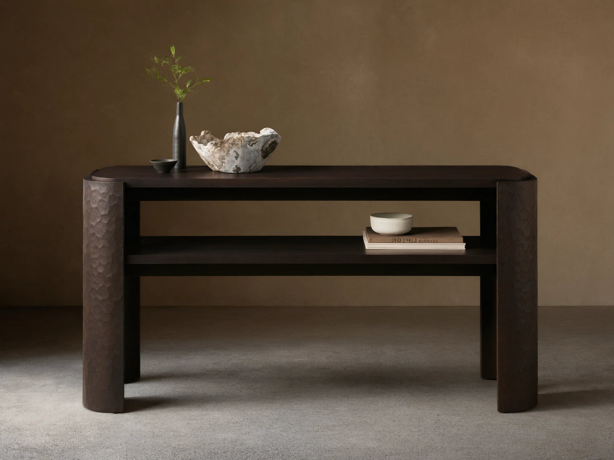 Dark brown mahogany Chiseled Console Table with rounded top, lower shelf. Legs feature distinct chiseled texture. Styled with decor.