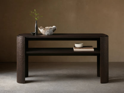 Dark brown mahogany Chiseled Console Table with rounded top, lower shelf. Legs feature distinct chiseled texture. Styled with decor.