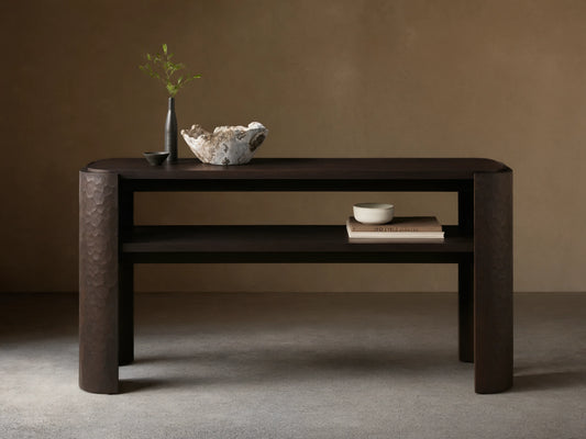 Dark brown mahogany Chiseled Console Table with rounded top, lower shelf. Legs feature distinct chiseled texture. Styled with decor.
