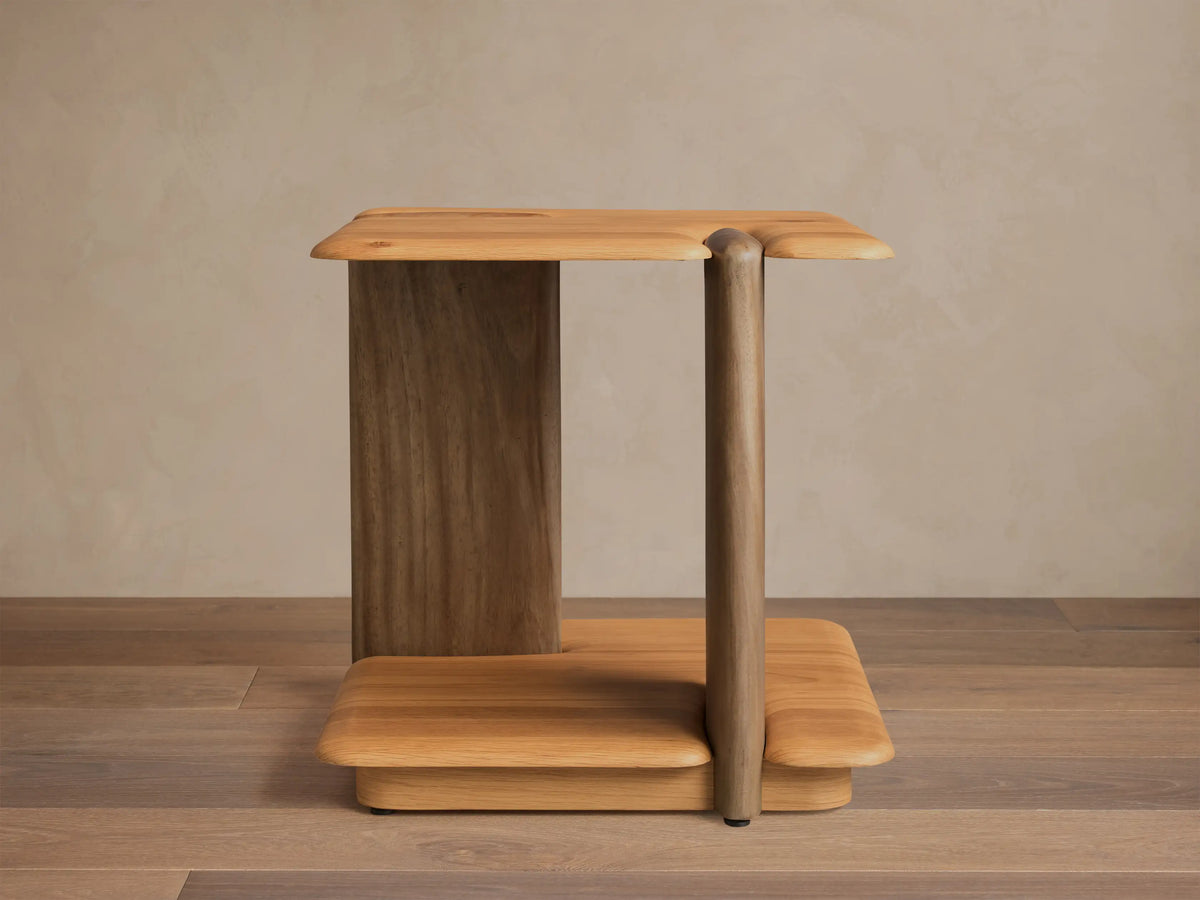 Front view of a light brown wooden side table, featuring a flat top and base with rounded edges, supported by a wide slab and a rounded leg.