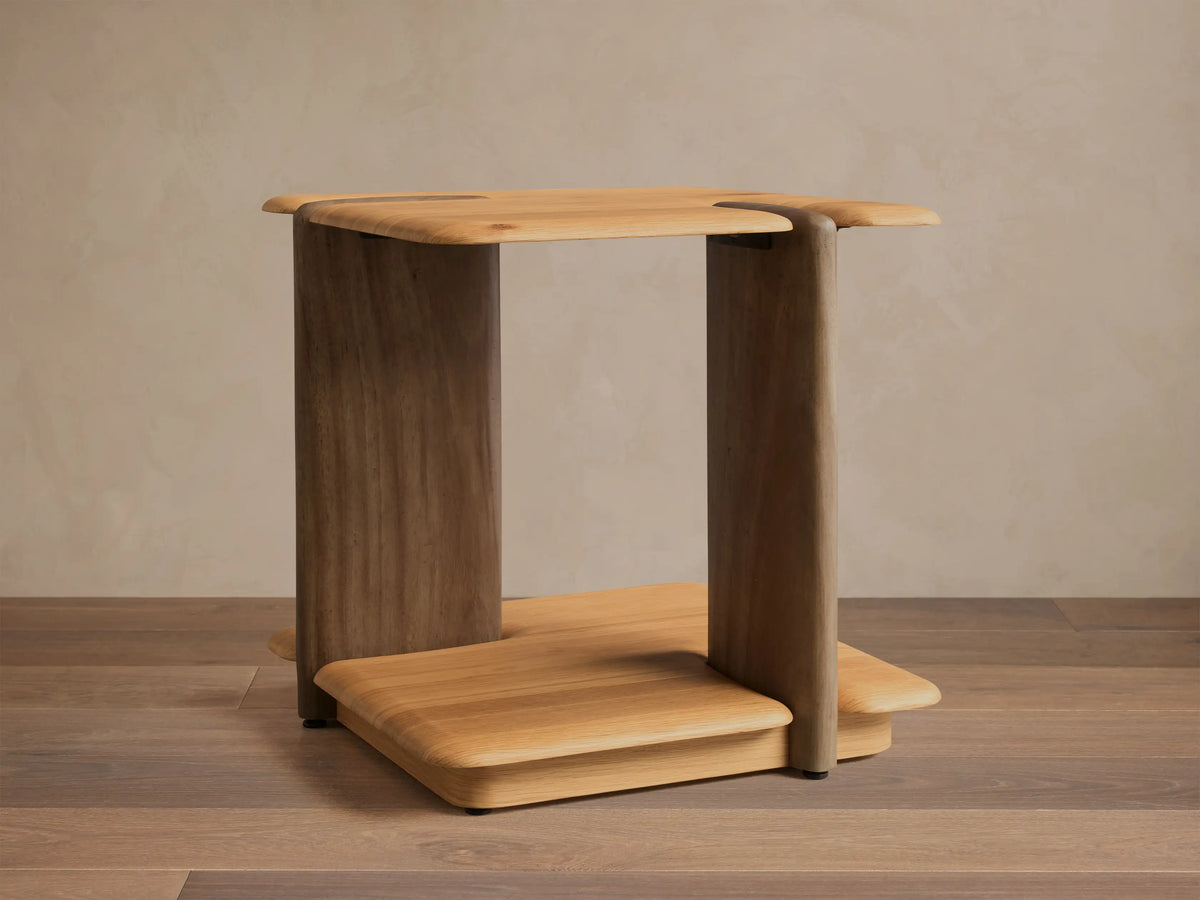 Front view of a light brown wooden side table, featuring a flat top and base with rounded edges, supported by a wide slab and a rounded leg.