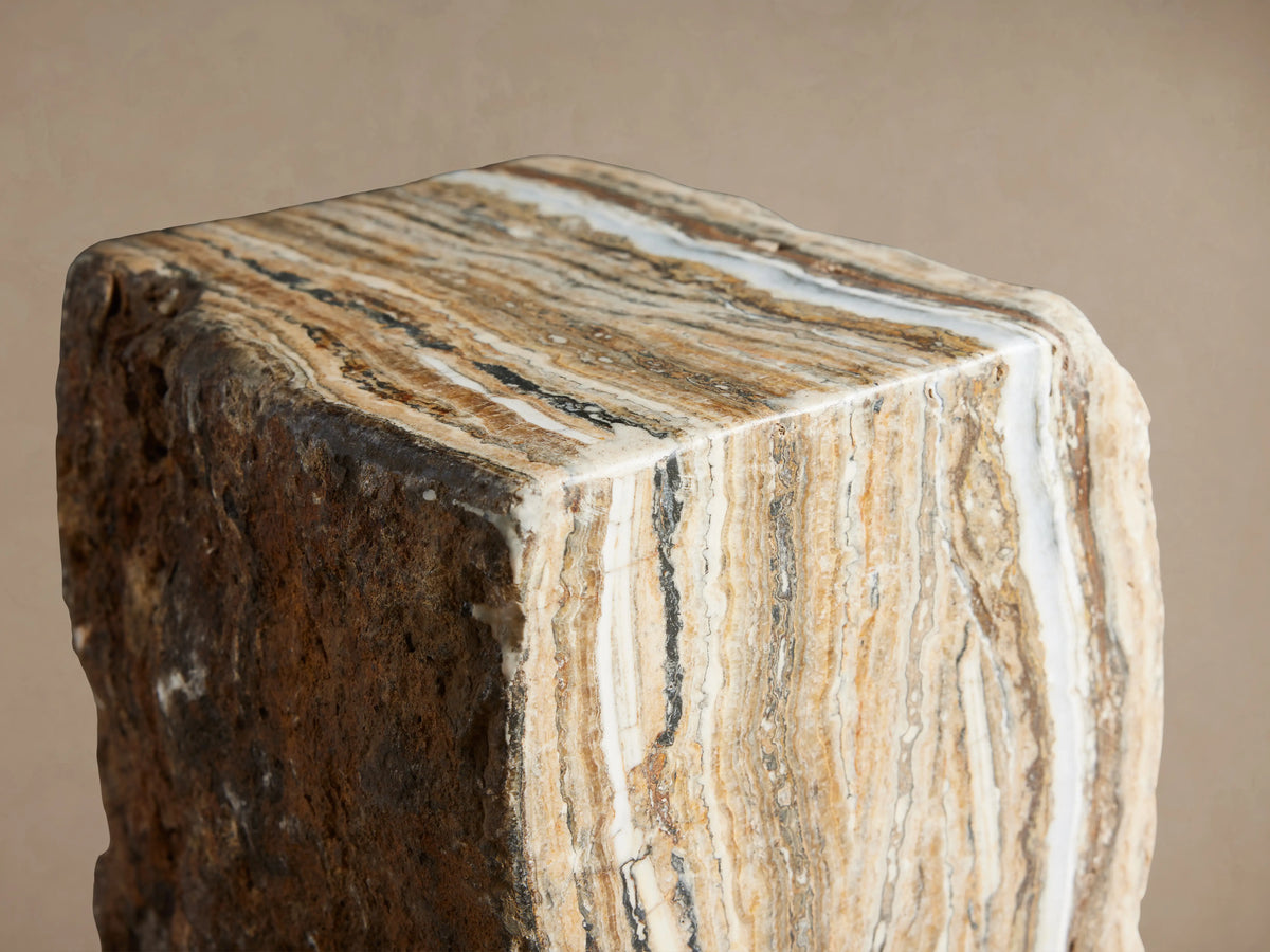 Solid Onyx Stool with vertical striations in warm brown and white tones, raw textured edges; stands on a wooden floor.