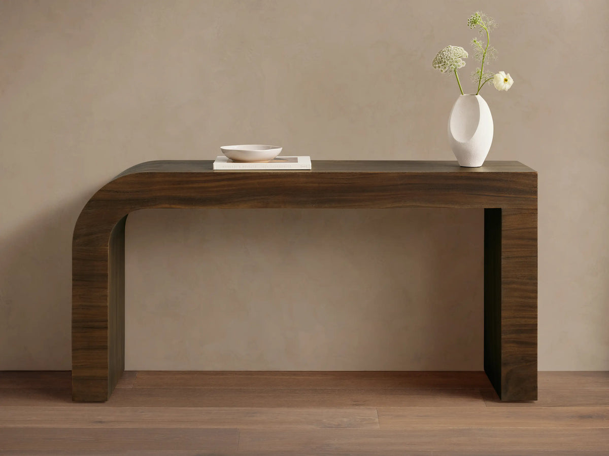 Dark brown Pliego Console Table with curved left leg. White vase with flowers and small bowl on top, against a beige wall.