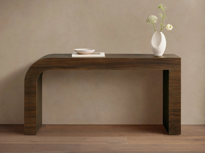 Dark brown Pliego Console Table with curved left leg. White vase with flowers and small bowl on top, against a beige wall.