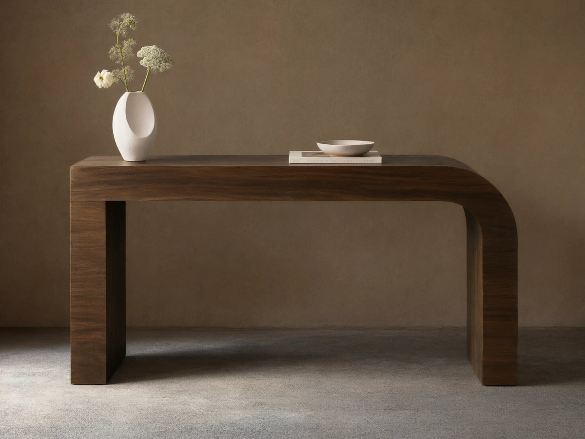 Dark brown Pliego Console Table with curved left leg. White vase with flowers and small bowl on top, against a beige wall.