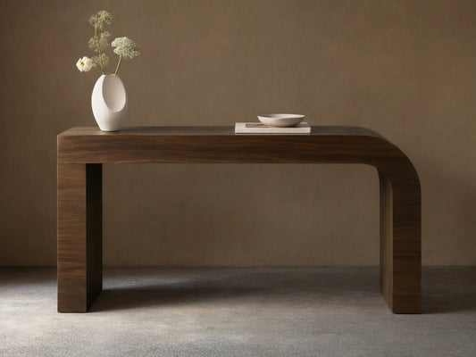 Dark brown Pliego Console Table with curved left leg. White vase with flowers and small bowl on top, against a beige wall.