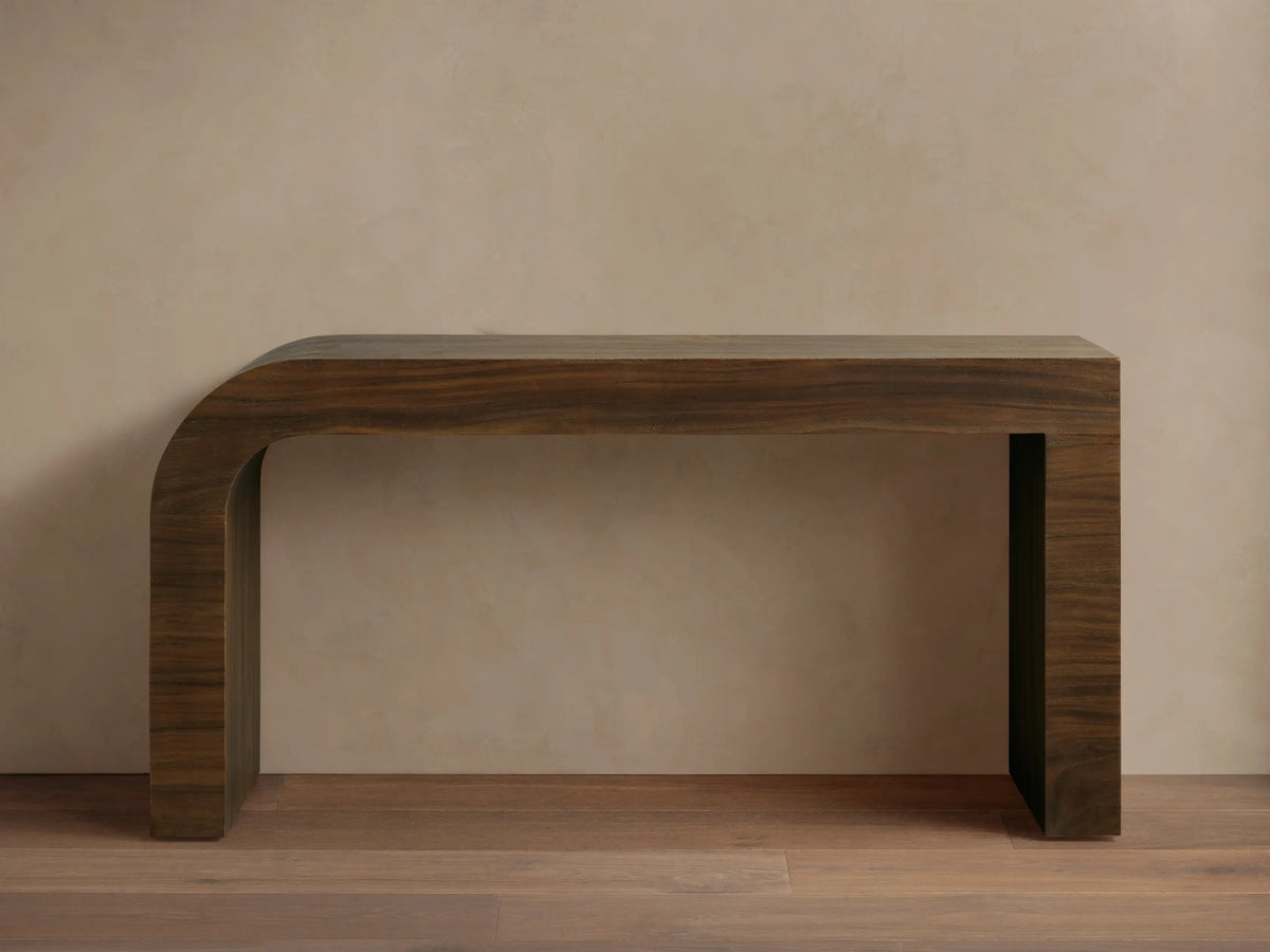 Dark brown Pliego Console Table with curved left leg. White vase with flowers and small bowl on top, against a beige wall.