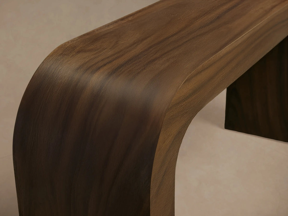 Pliego Console Table, dark wood with waterfall design on left, straight leg on right, against a neutral wall on wood floor.