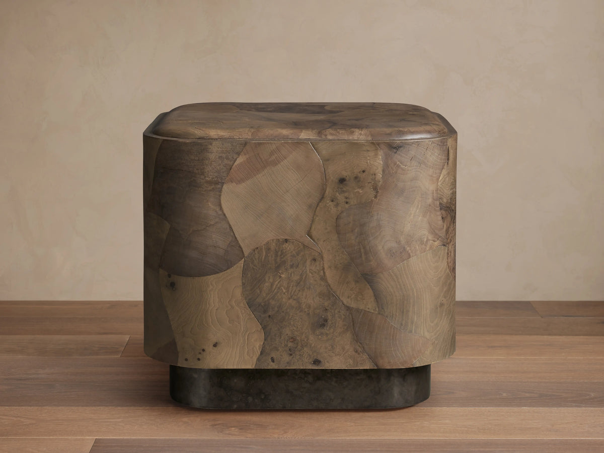 Polanco Plinth End Table, a block-shaped table with rounded edges, crafted from varied brown oyster-cut wood burl fragments. Dark plinth base.