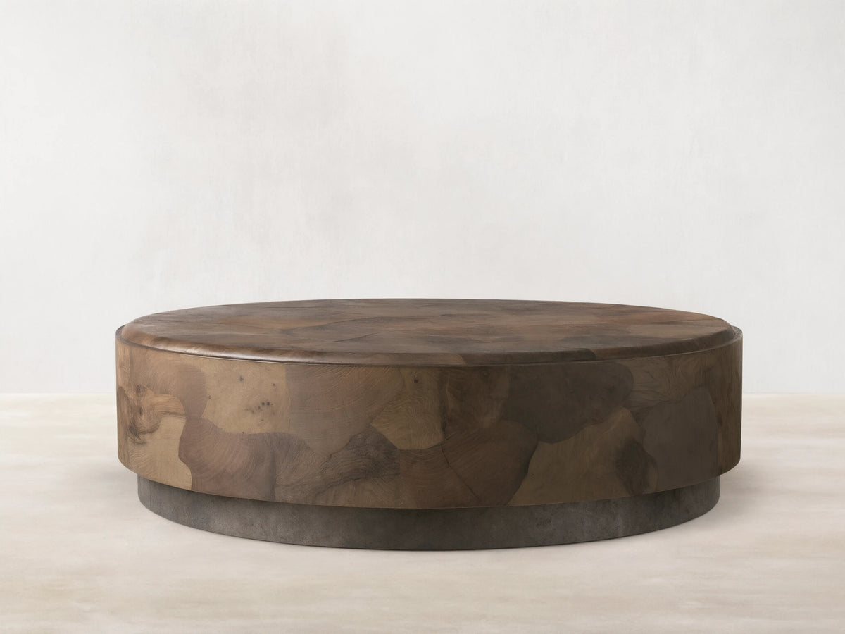 Round coffee table with smooth, curved edges and a textured surface. Made from ash wood fragments, showing tonal variations and natural patterns. Set on a weathered steel base with rich patina.