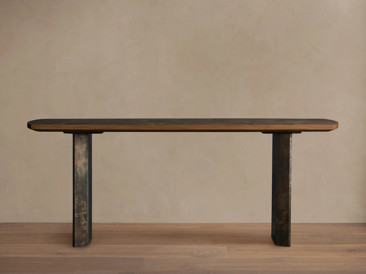 Polanco Console Table featuring an elongated wooden top with visible grain, supported by two steel legs with a weathered patina finish.