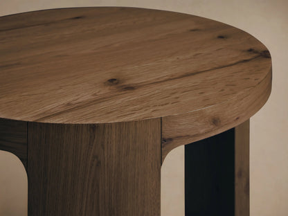 Front view of the round, light brown Redding End Table, featuring a built-in lower shelf and thick vertical supports.