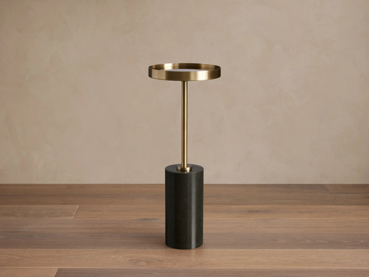 Rhodes Martini Table with a brass tray atop a slim stem and a black cylindrical marble base, set on a wooden floor.