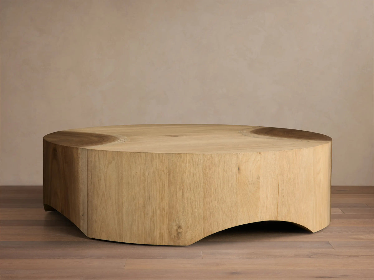Front view of a light wood coffee table, round with a solid, inwardly curved base, showcasing subtle top surface details.