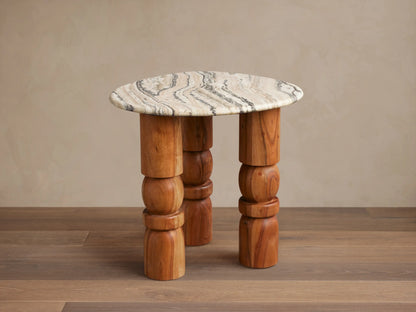 Rune End Table with a round, marbled stone top and unique carved wooden legs, set on a wooden floor against a beige wall.