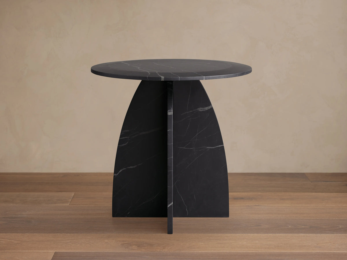 Round Solstice End Table made of Black Marquina marble with a smooth surface, curved legs, and wooden floor background.