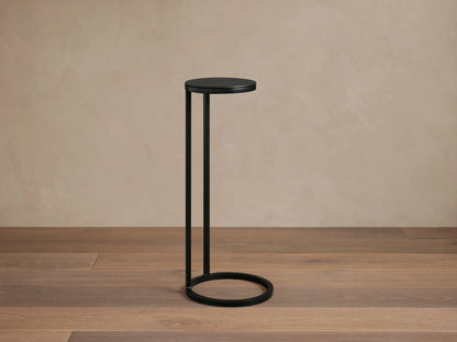Soma Martini Table with a sleek black metal frame, round top, and circular base, standing on a wooden floor against a beige wall.