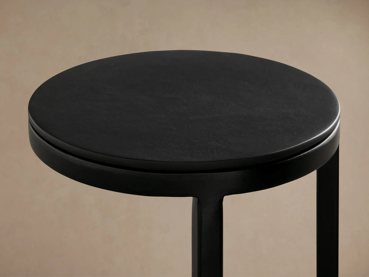 Soma Martini Table with a sleek black metal frame, round top, and circular base, standing on a wooden floor against a beige wall.