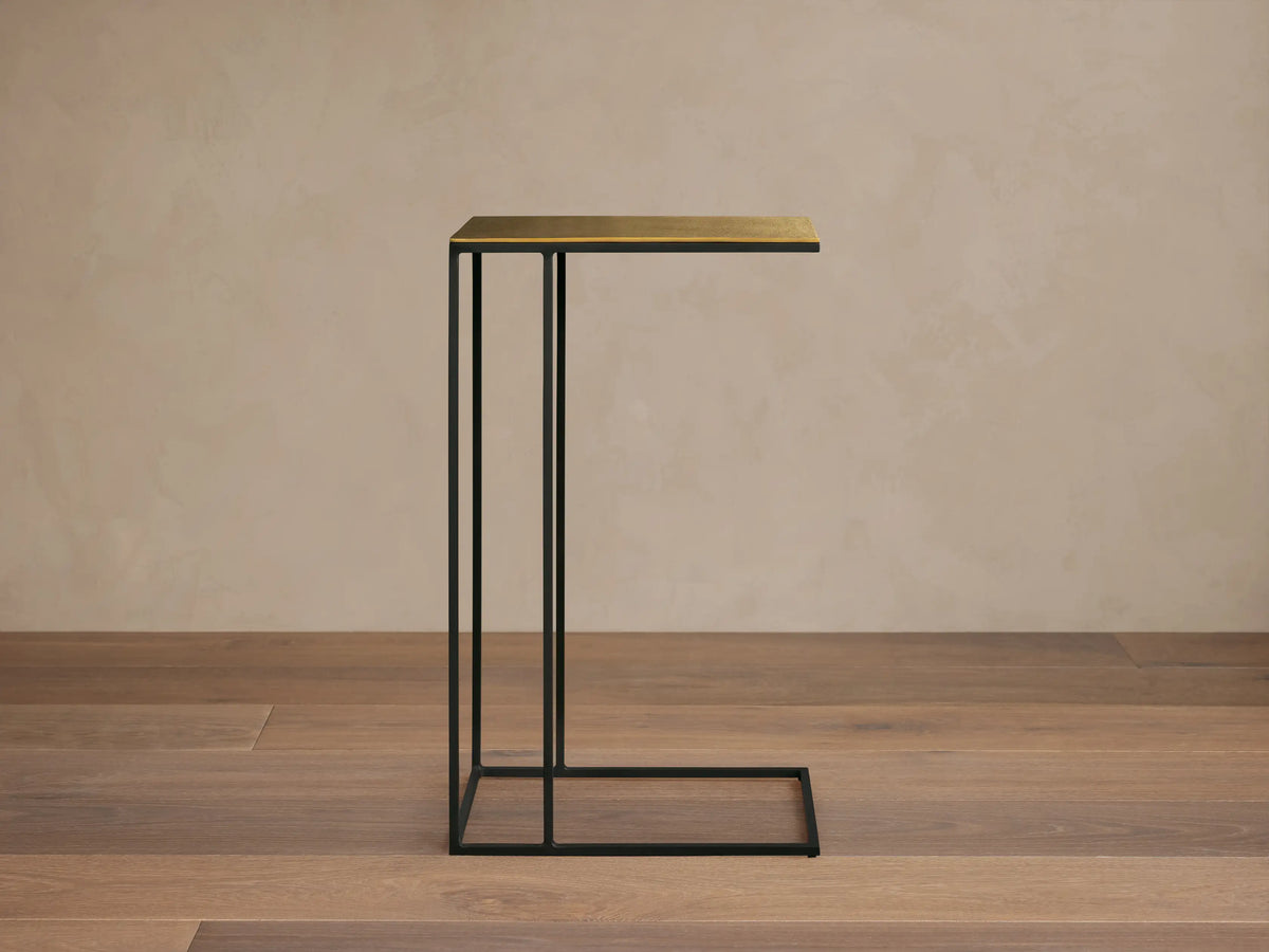 Front view of the Soma C Table, featuring a black C-shaped metal base and a rectangular gold-toned top.