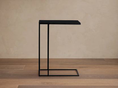 Black Soma C Table with dark rectangular top on C-shaped metal frame. Stands on textured light tiled floor against a pale wall.