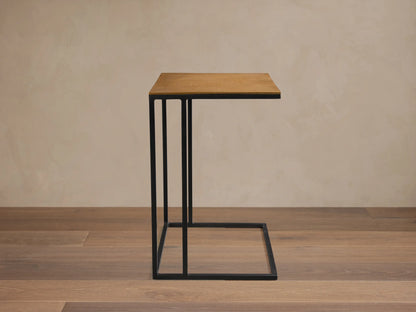 Soma Wide C Table with light brown wooden top and black metal C-frame, on a stone floor against a beige wall.