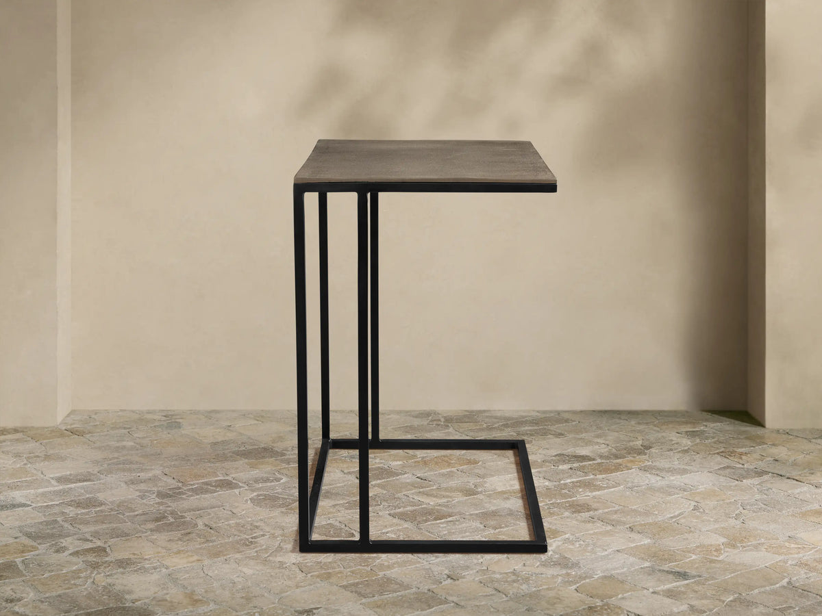 Soma Wide C Table, dark gray patinaed rectangular top, black powder-coated C-shaped base on a textured stone floor.