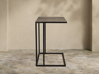 Soma Wide C Table, dark gray patinaed rectangular top, black powder-coated C-shaped base on a textured stone floor.