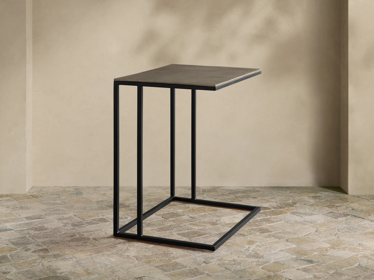 Soma Wide C Table, dark gray patinaed rectangular top, black powder-coated C-shaped base on a textured stone floor.