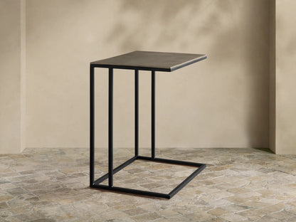 Soma Wide C Table, dark gray patinaed rectangular top, black powder-coated C-shaped base on a textured stone floor.