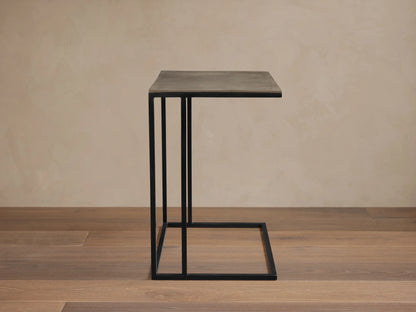 Soma Wide C Table, gray square top on black powder-coated C-shaped base, angled view on light tiled floor against tan wall.