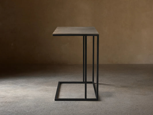 Soma Wide C Table, dark gray patinaed rectangular top, black powder-coated C-shaped base on a textured stone floor.