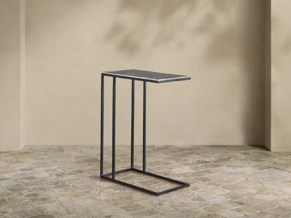 Soma C Table with a dark gray rectangular top and black C-shaped metal base, against a beige wall on a stone patio.