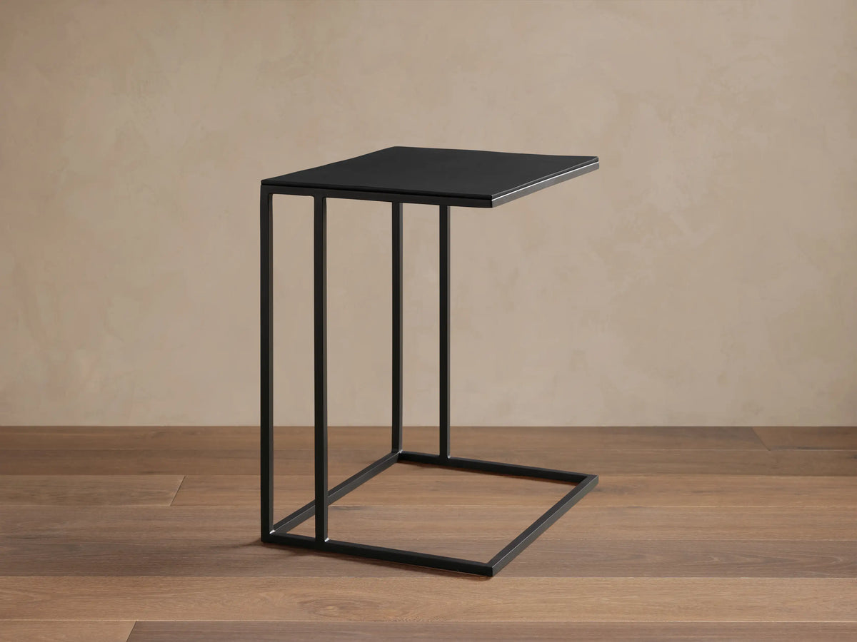 Black Soma C Table with square top and slender C-shaped metal frame on a wooden floor against a light wall.