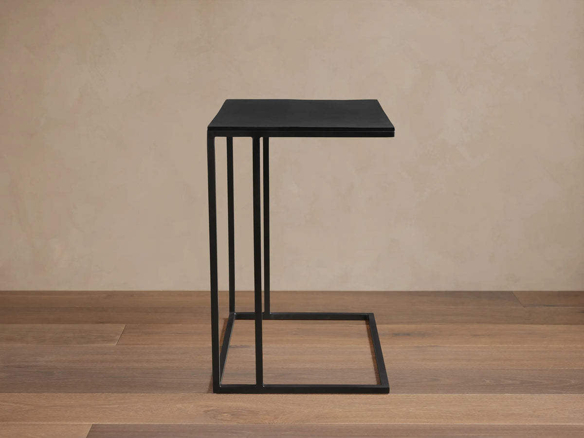 Black Soma C Table with a square top and slim metal frame, standing on a wooden floor against a light beige wall.