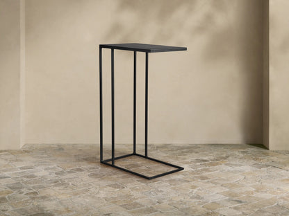 Black Soma C Table, featuring a rectangular top and sleek C-shaped base, standing on a textured tiled floor in a neutral room.