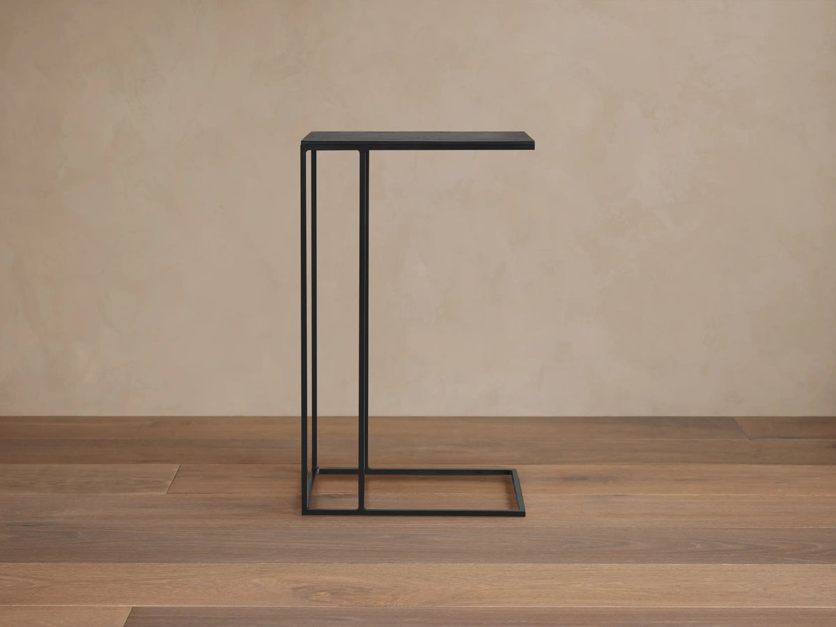 Soma C Table: Modern black metal C-shaped table with a square top, on a stone floor against a beige wall.
