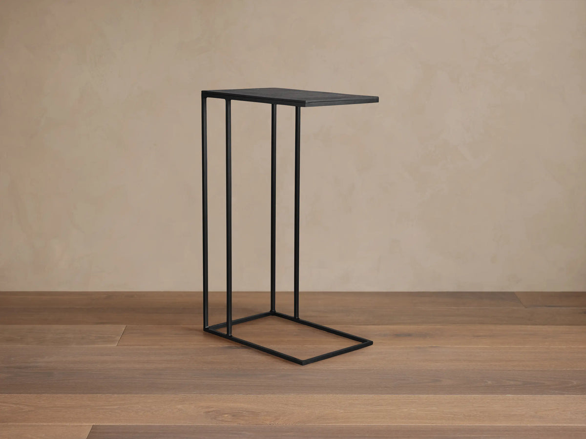 Black metal Soma C Table with a slender C-shaped frame on a wooden floor against a textured beige wall.
