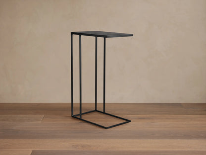 Black metal Soma C Table with a slender C-shaped frame on a wooden floor against a textured beige wall.