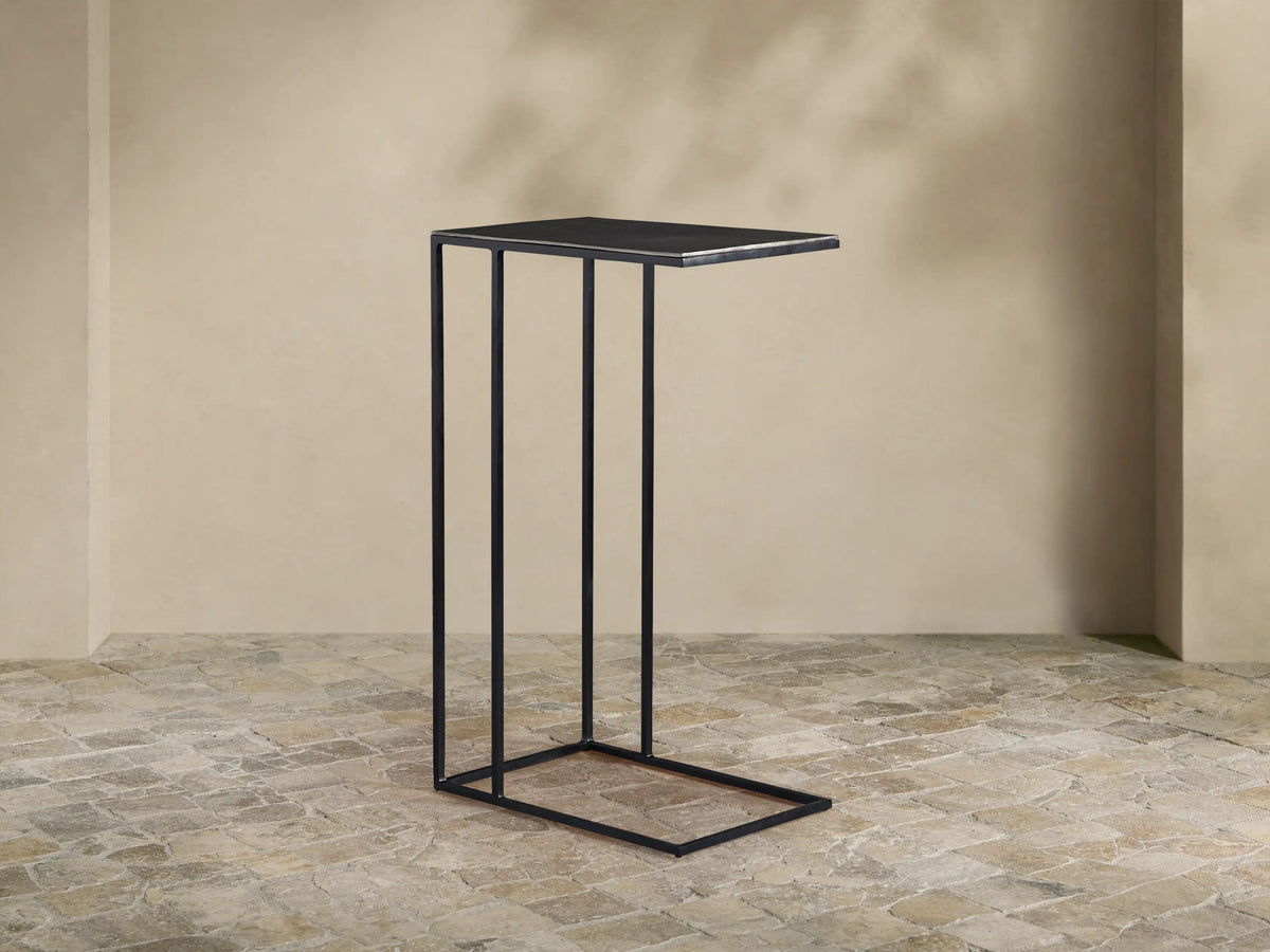 Black Soma C Table with a rectangular dark tabletop and thin metal C-frame, on a light stone floor against a beige wall.
