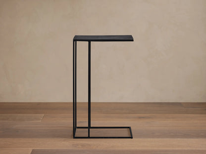 Soma C Table: a black C-shaped metal side table with a dark, rectangular tabletop, standing on a light stone floor.
