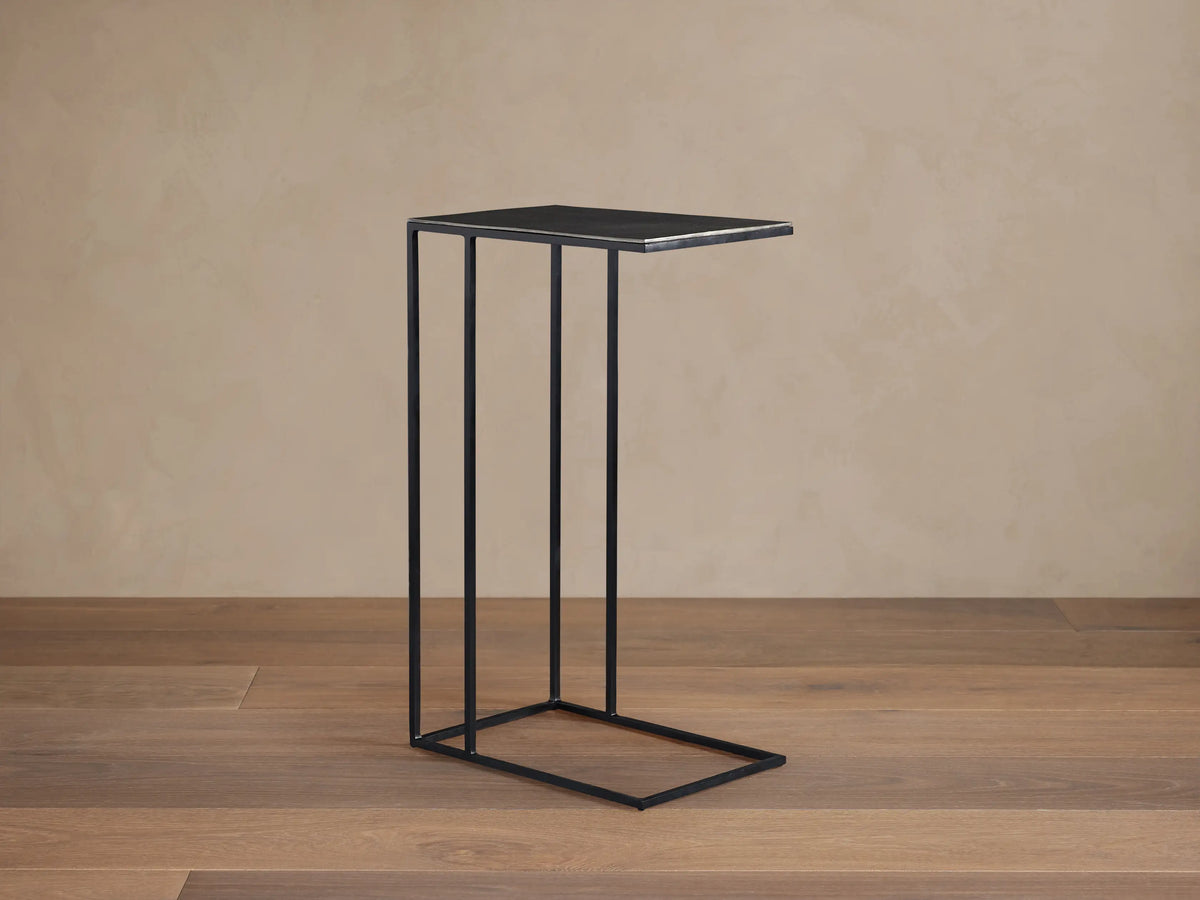 Black Soma C Table; metal C-shaped frame with a rectangular top and open base, on a wooden floor against a textured beige wall.