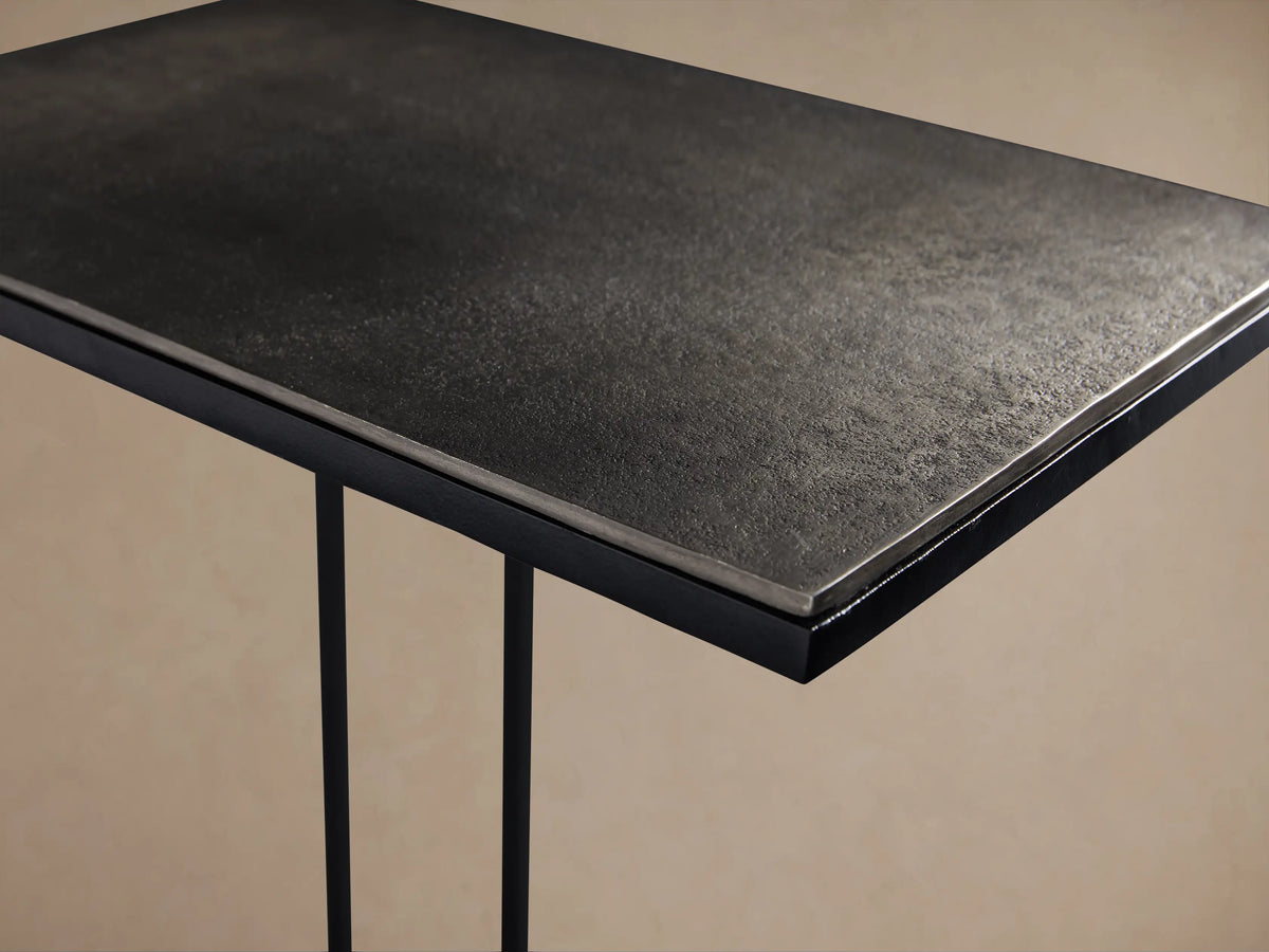 Soma C Table with black C-shaped metal frame & dark rectangular aluminum top, on wooden floor against light wall.