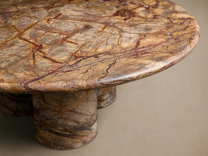 Front view of the round Serafin Coffee Table made from brown and reddish-brown marble with intricate veining and three cylindrical legs.