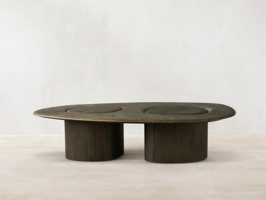Sucurro Coffee Table: Elongated oxidized aluminum top with two dark circular depressions, supported by two thick dark wood bases.